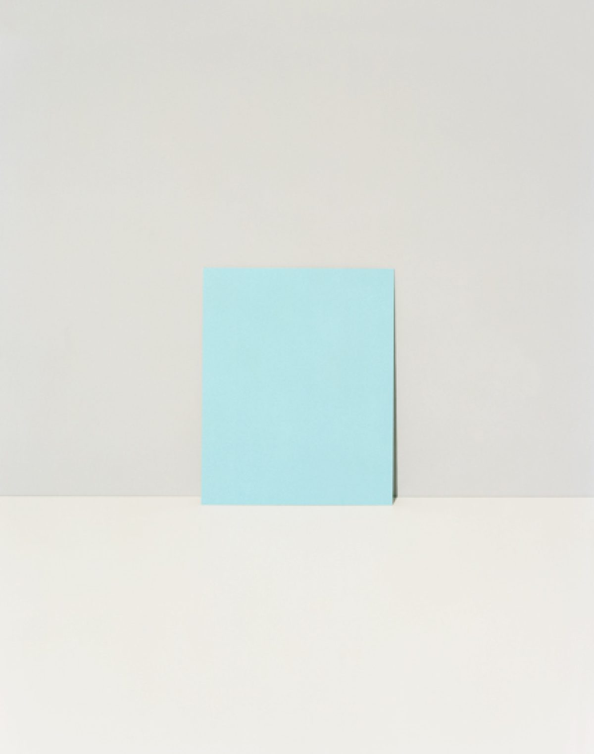 A light blue rectangular form stands vertically against a neutral beige wall and floor, casting a subtle shadow. The minimalist composition emphasizes the geometric shape and its interaction with the surrounding space.