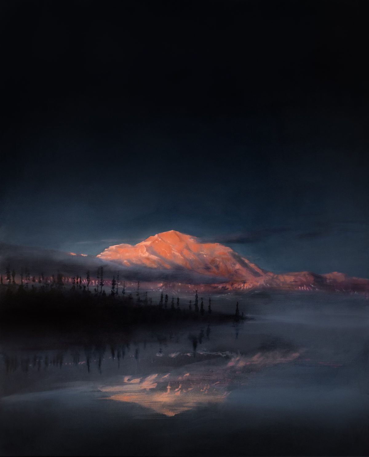 A snow-capped mountain peak glows in warm amber and orange tones against a deep blue twilight sky, its reflection mirrored in still water below. Silhouetted evergreen trees line the shoreline in the foreground, creating a stark contrast between the illuminated summit and the dark landscape.