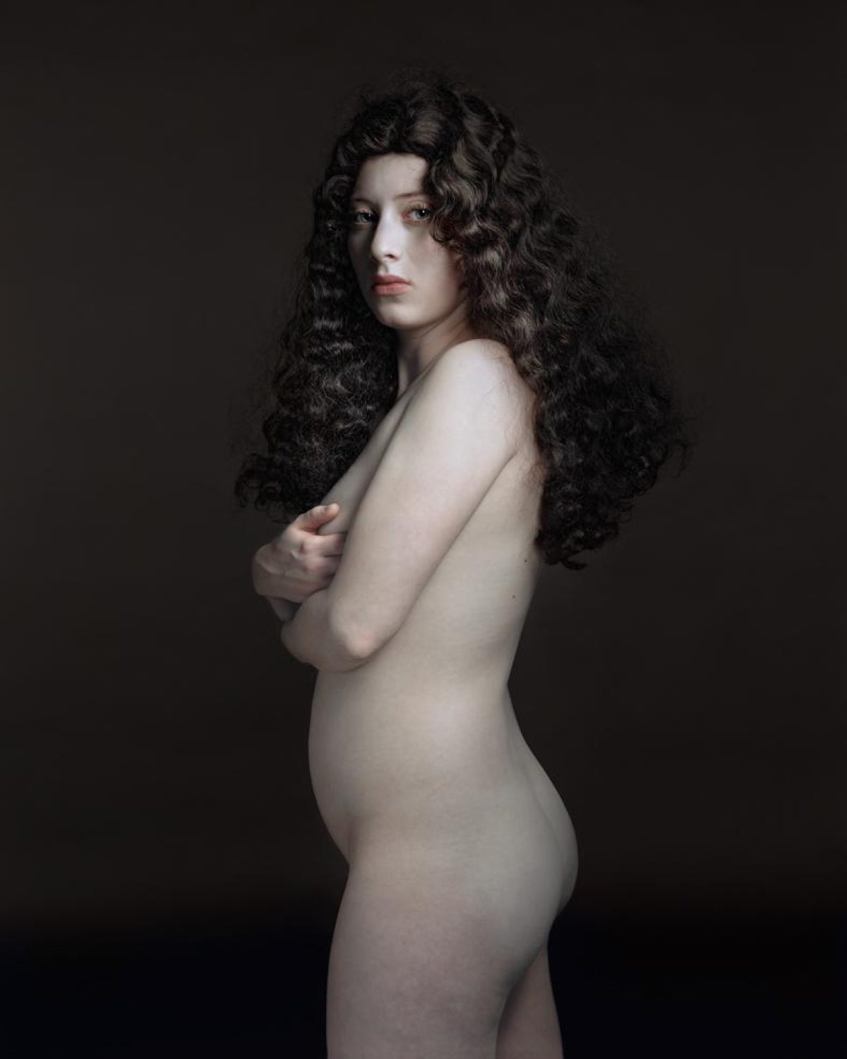 Portrait of a woman with long, voluminous dark curly hair shown in profile against a dark neutral background. She is posed with her body turned away and arm crossed, rendered in pale tones that create strong contrast with the shadowed backdrop.