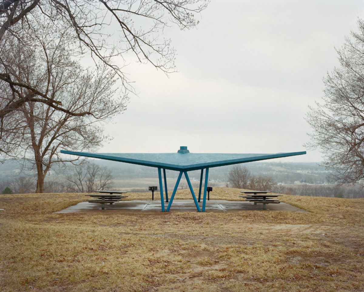 A large teal-blue sculptural shelter with an expansive wing-like roof structure stands in a sparse winter landscape. Bare trees frame the composition on either side, with picnic tables positioned beneath the canopy and rolling hills visible in the distant background.