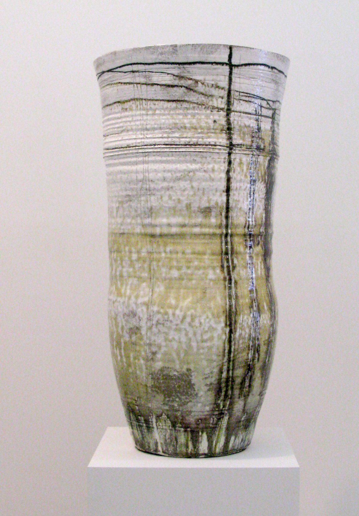 A tall cylindrical stoneware vessel with a matte surface featuring horizontal linear markings and vertical incised lines. The vessel transitions in color from pale gray-white at the rim to muted green and ochre tones toward the base, with darker accretions suggesting natural weathering or firing effects.