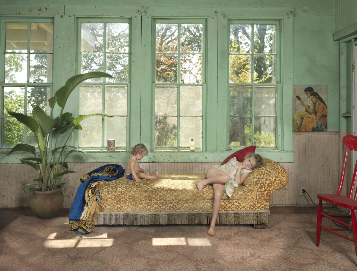 Two children play and rest on a patterned daybed in a sunlit room with mint-green walls and large gridded windows overlooking trees. A potted plant, red chair, and framed artwork are visible in the softly lit interior space.