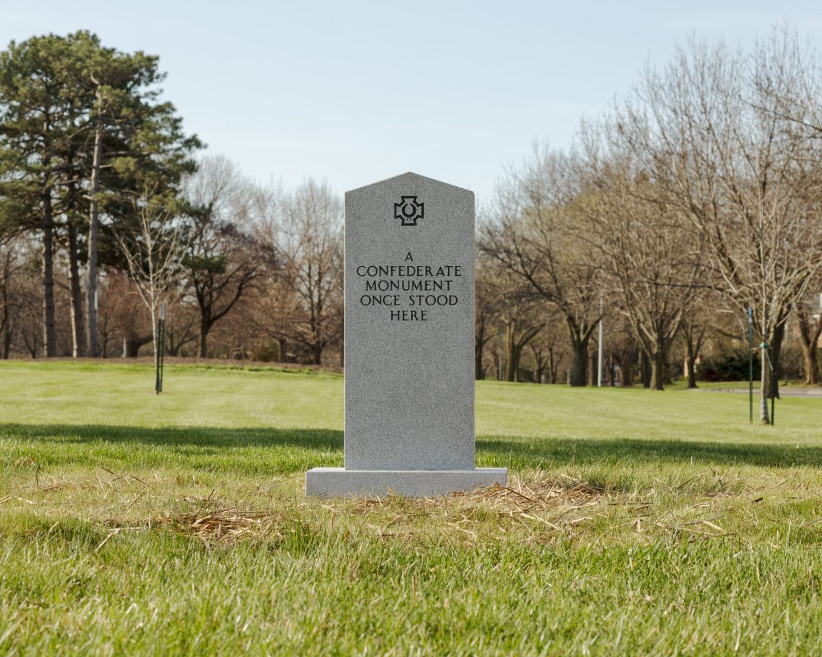 A gray stone monument stands alone in a grassy park surrounded by bare and evergreen trees. The obelisk-shaped marker bears an engraved symbol at its top and text reading "A CONFEDERATE MONUMENT ONCE STOOD HERE."