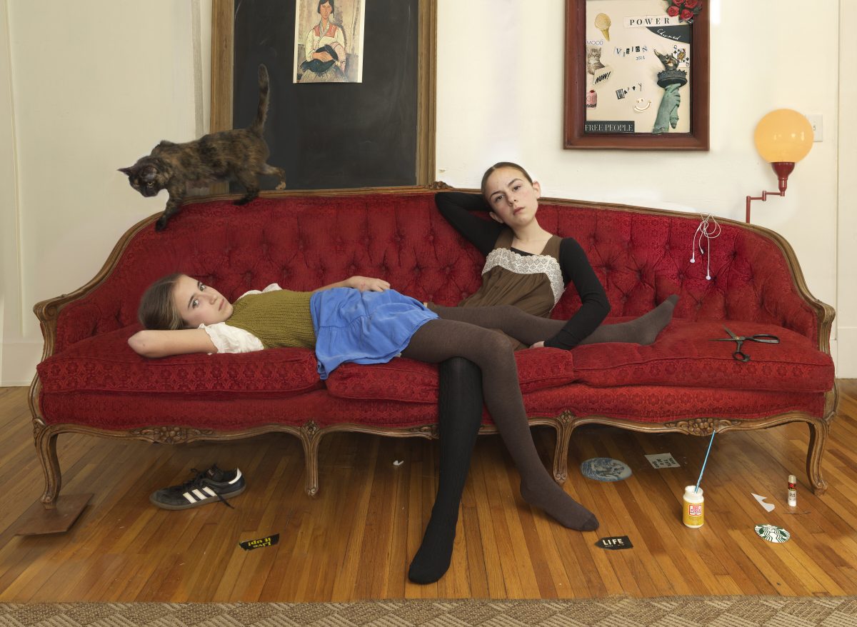 Two young women recline on a red velvet sofa with ornate wooden frame in a domestic interior. One lies across the cushions while the other sits upright; a tabby cat walks along the back. The wooden floor is scattered with magazines, scissors, and other craft materials. Framed artworks and a yellow wall-mounted lamp are visible in the background.