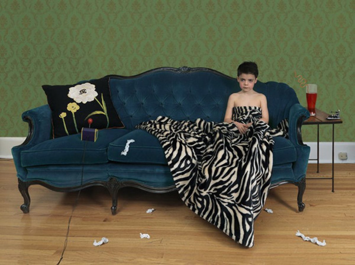 A person in a black strapless top reclines on a teal upholstered sofa draped with a zebra-striped blanket, against a green damask wallpapered wall. Scattered tissues and objects lie on the wooden floor, while a side table with a glass holds personal items nearby, creating a scene of domestic illness or malaise.