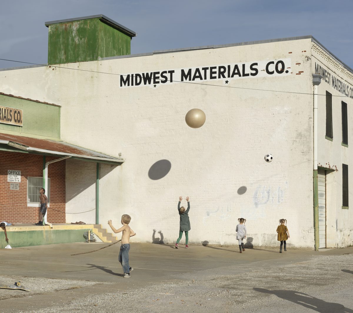 Children play in a sunny parking lot in front of a weathered cream-colored industrial building with "Midwest Materials Co." painted across its facade. Painted spheres and a soccer ball adorn the blank wall, transforming the utilitarian structure into an imaginative play space.
