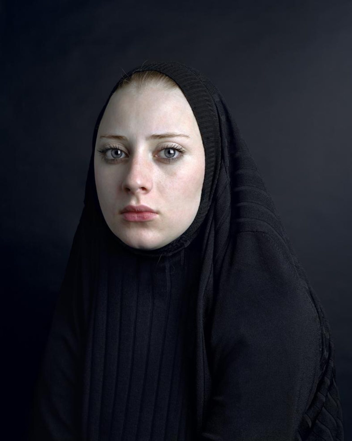 Portrait of a pale-skinned woman with blue eyes gazing directly at the camera, her head covered with a black textured fabric draped over her shoulders against a dark gray background. The lighting is stark and frontal, emphasizing the contrast between her fair complexion and the dark surrounding fabric.