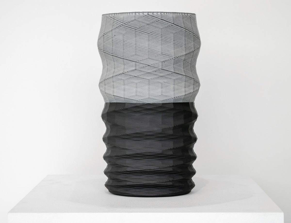 A tall cylindrical vessel with a twisted, ribbed surface sits on a white platform. The upper half features a translucent gray mesh-like texture, while the lower half is rendered in solid black with horizontal ridging, creating a striking color and material contrast.