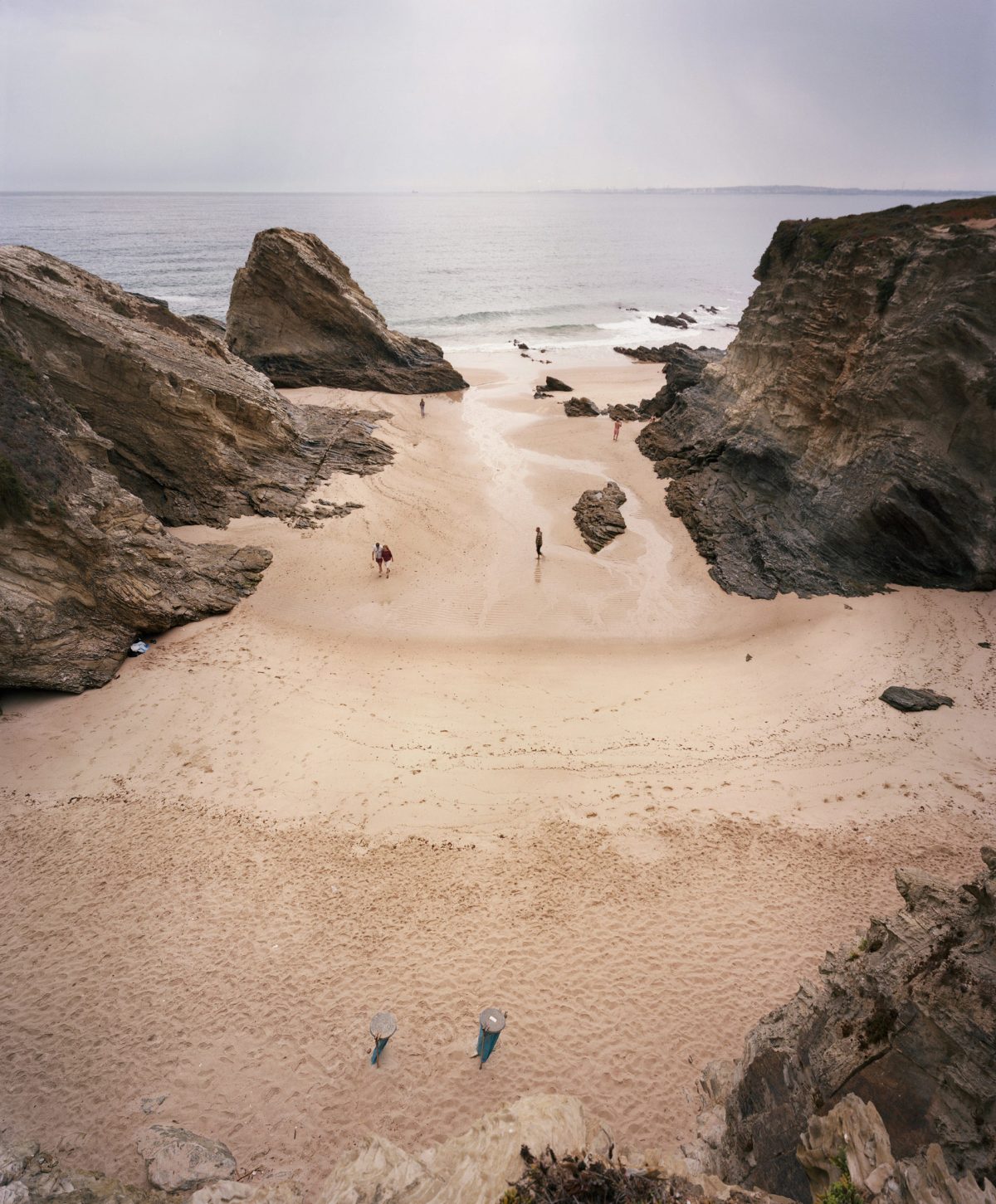 A secluded sandy beach enclosed by dramatic rocky outcrops and large boulders, with several small figures scattered across the sand and wading in shallow water. The composition is framed by weathered stone formations in the foreground and middle ground, while calm ocean and pale sky occupy the background.