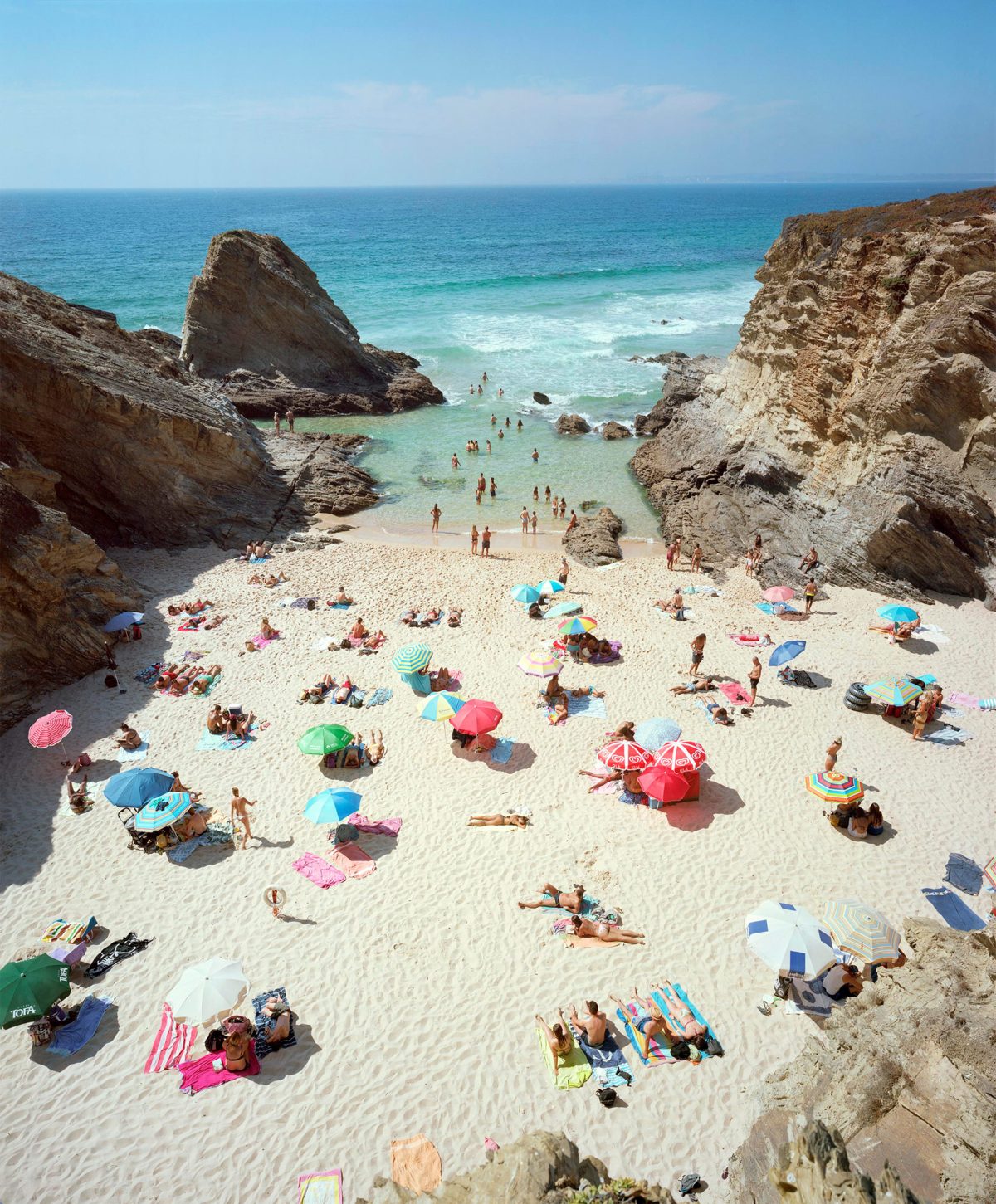 Aerial view of a crowded sandy beach nestled between dramatic limestone cliffs, with colorful umbrellas and sunbathers scattered across the pale sand, turquoise shallow waters filled with swimmers, and deeper blue ocean beyond.