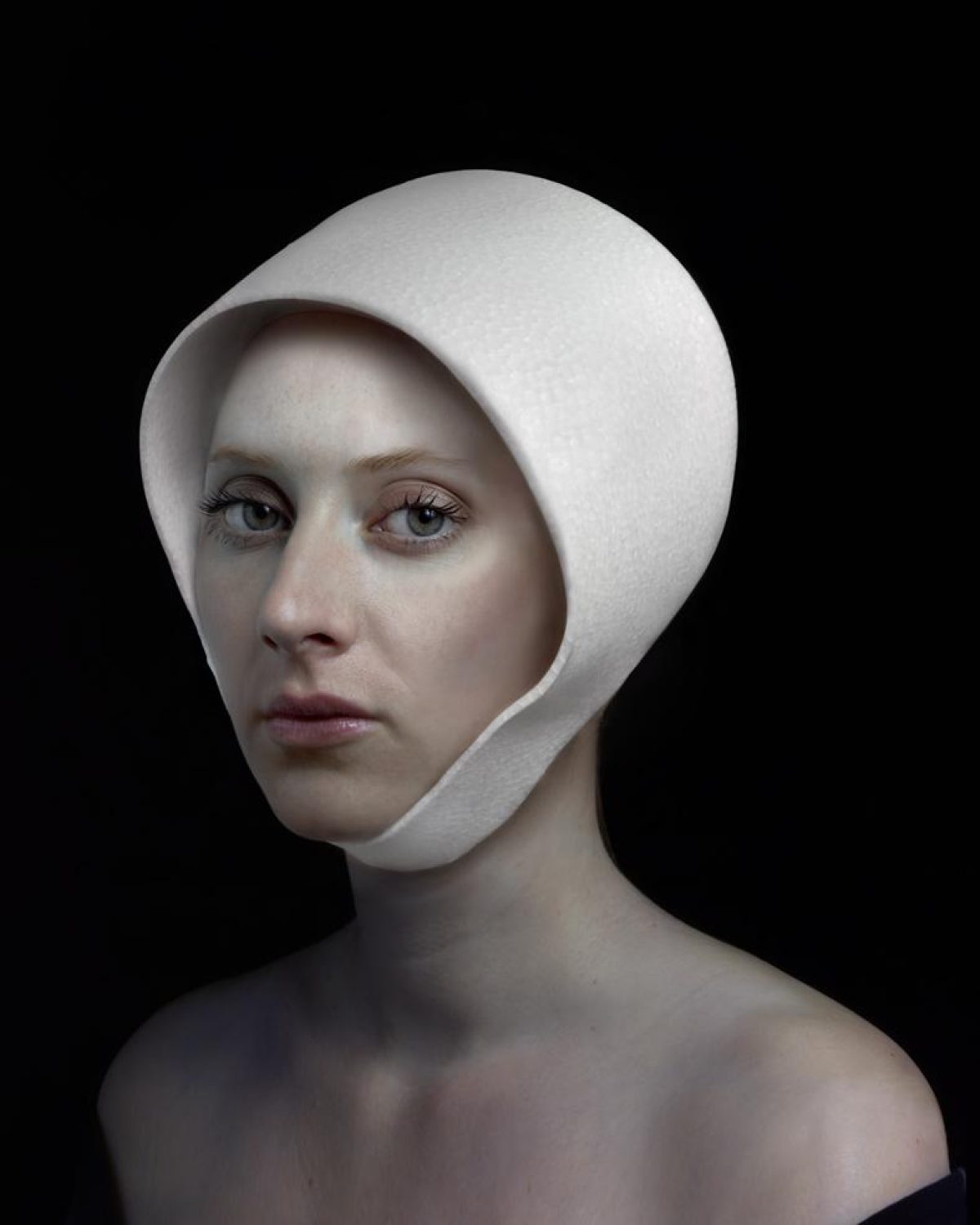 Portrait of a pale-skinned woman with dark eyes and lips against a black background, her head wrapped in a white fabric hood that frames her face. The minimalist composition emphasizes the subject's contemplative expression and the stark contrast between her skin tone and the pristine white textile.