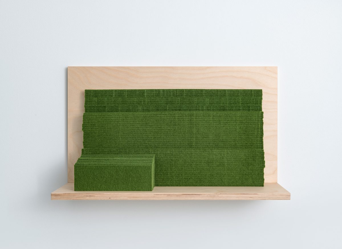 A wooden shelf mounted on a white wall holds two green felt rectangular forms of different sizes—a smaller one positioned on the left and a larger one behind it—creating a minimalist sculptural composition.
