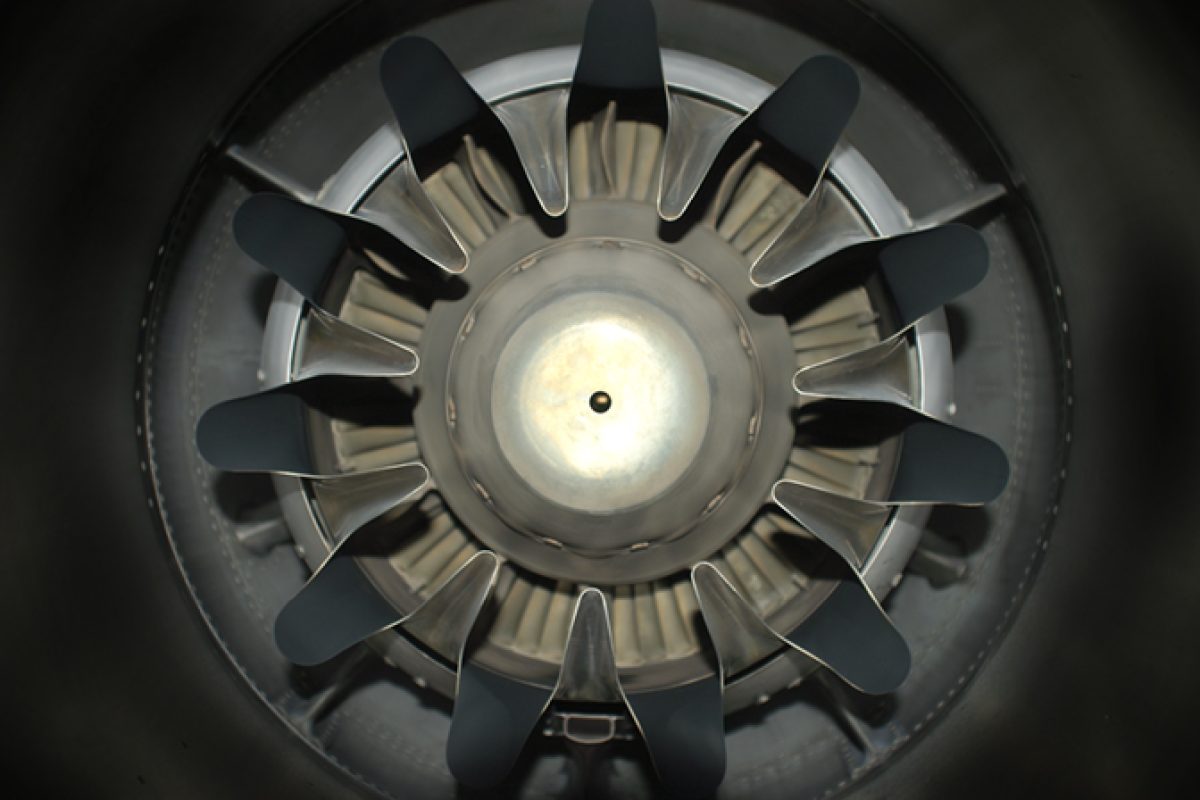 Overhead view of a circular mechanical structure with black blade-like forms radiating outward from a pale central hub, photographed against a dark background. The composition emphasizes radial symmetry and industrial materiality, with metallic and matte surfaces creating subtle tonal contrasts.