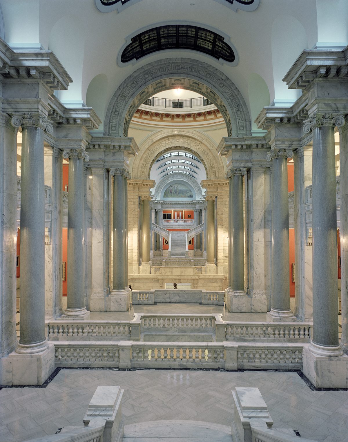 A symmetrical corridor with towering fluted columns in pale marble flanks a central passage, leading to a grand arched rotunda with ornamental detailing. Warm lighting illuminates the neoclassical architecture, while burnt orange accent panels and ornate balustrades recede into the distance.