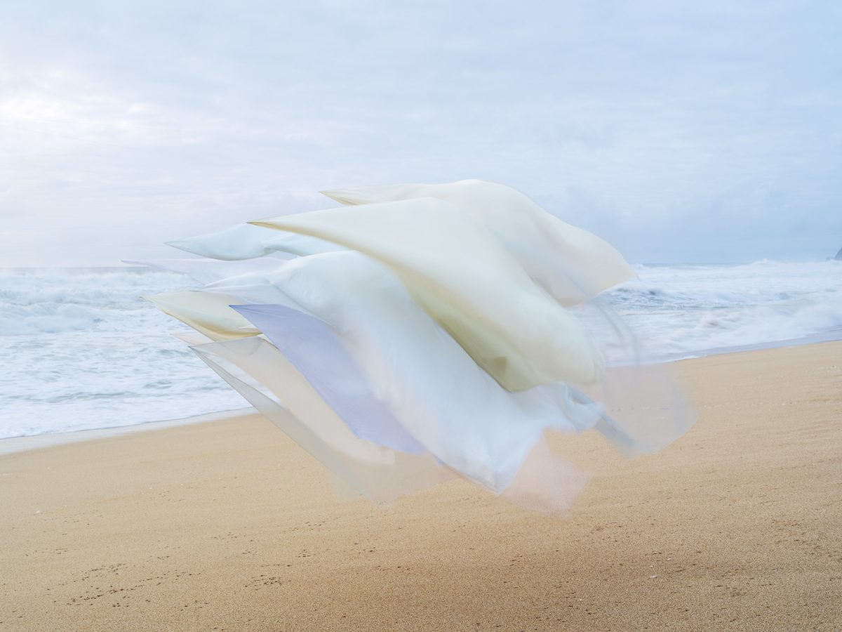White and cream-colored silk fabric billows and folds across a sandy beach, with ocean waves and overcast sky visible in the background. The translucent material catches subtle shadows of blue and lavender as it moves in the wind.