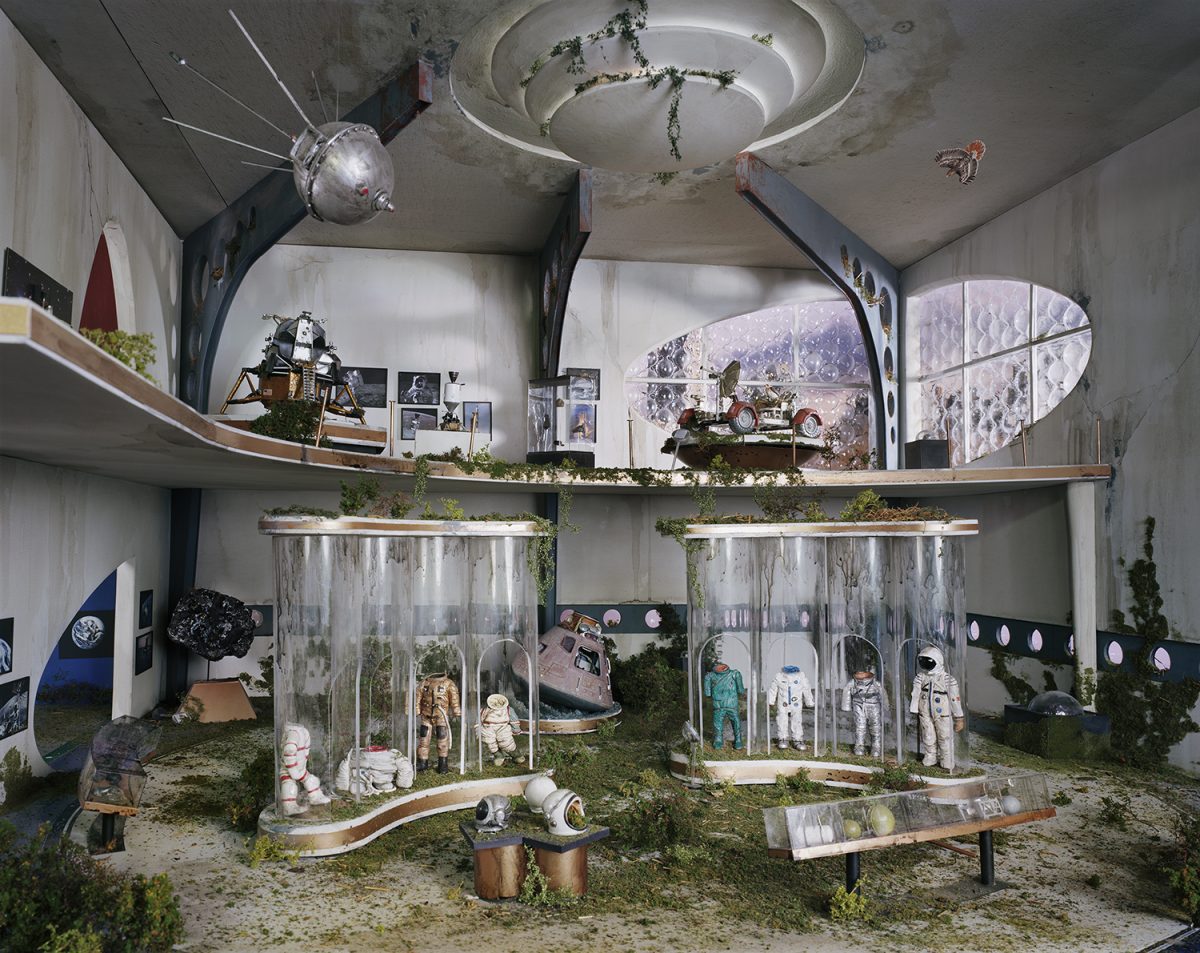 An abandoned institutional interior overgrown with vegetation features glass display cases containing astronaut suits and space equipment, with a Soyuz capsule and lunar rover visible on upper shelves. Concrete walls, debris-strewn floors, and celestial imagery suggest a decaying space museum frozen in time.
