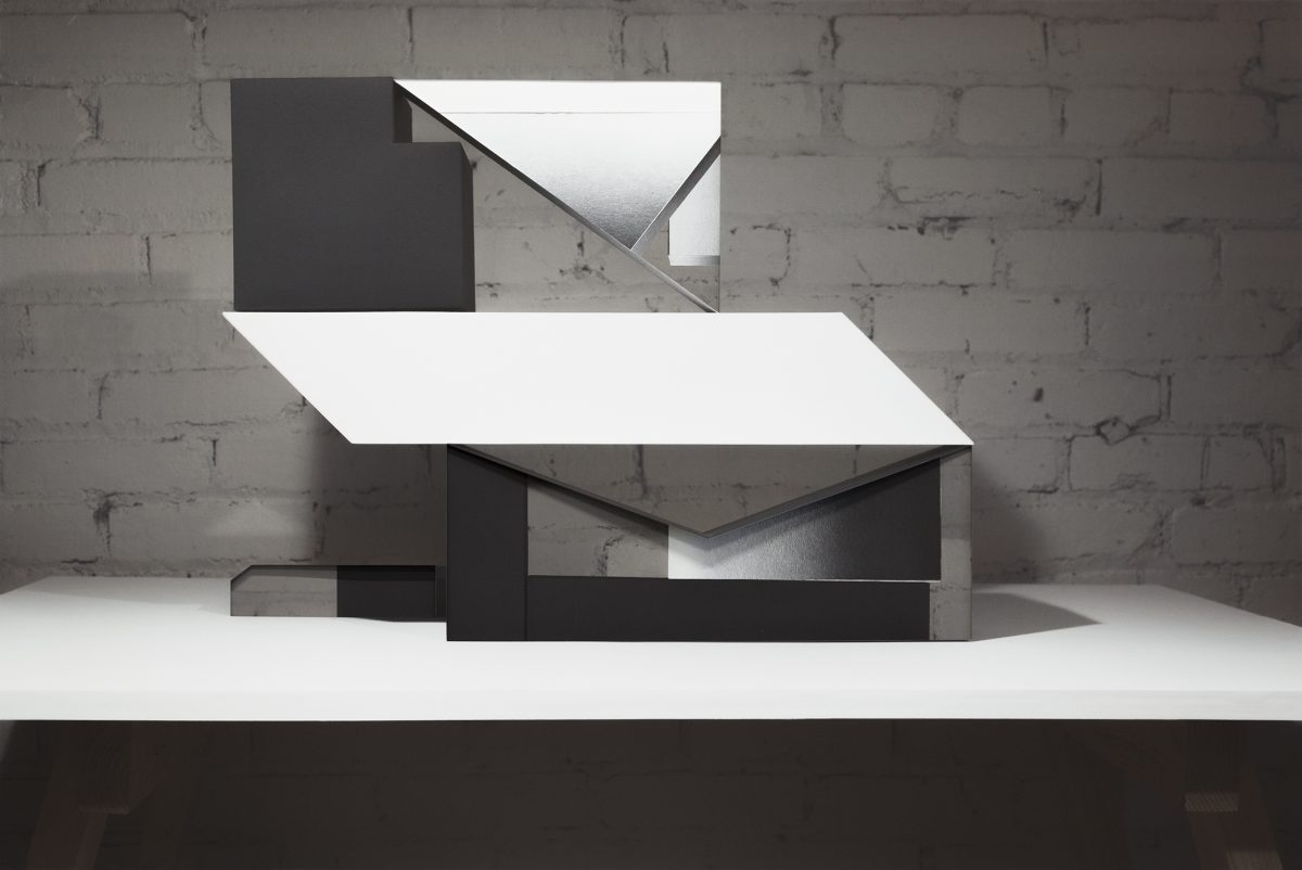 An abstract three-dimensional sculptural form constructed from layered white and black materials, displayed on a white platform against a weathered brick wall. The composition features angular, intersecting planes with folded and cut surfaces that create dynamic shadows and spatial depth.