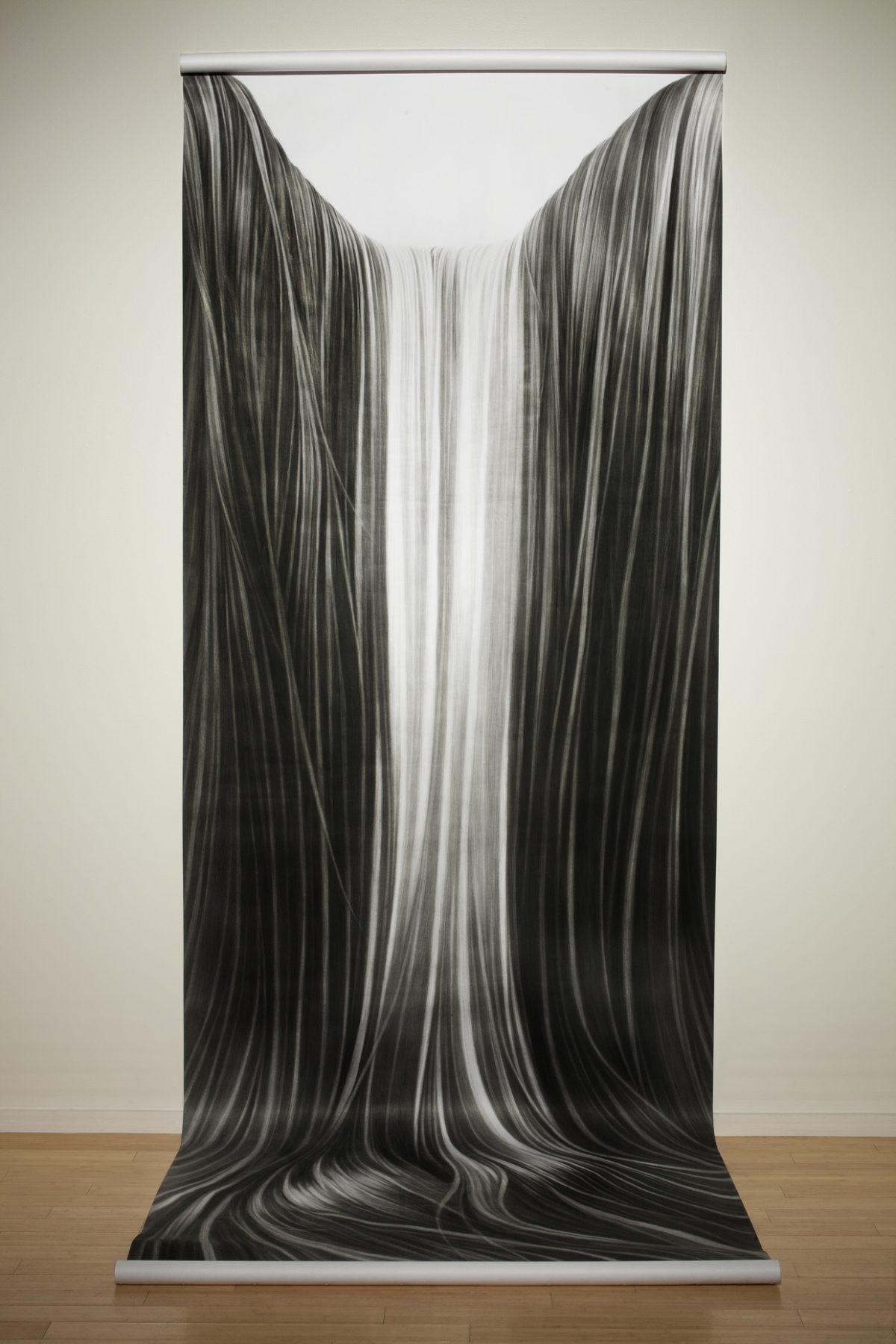 A monumental vertical drawing in charcoal depicts flowing, hair-like strands cascading downward in sinuous curves. The composition features darker tones at the edges that gradually lighten toward a luminous central channel, creating a sense of movement and depth as the linear forms pool and gather at the base.