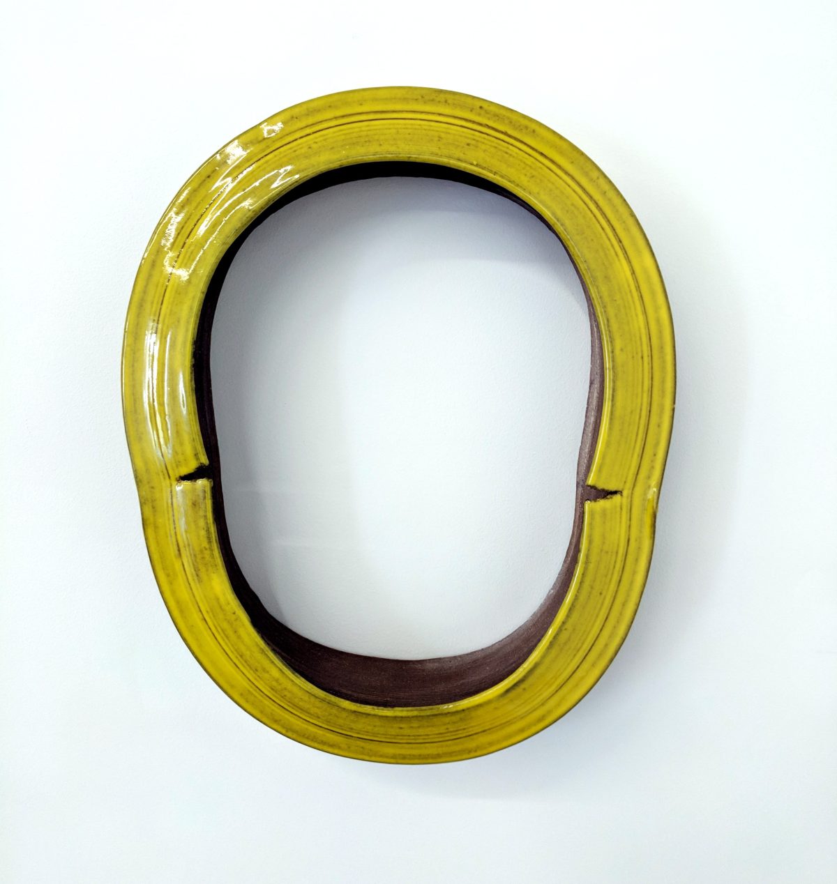 A vibrant yellow-green glazed ceramic ring form with an oval opening, set against a white background. The glossy surface contrasts with the darker interior, and subtle linear textures run across the frame's width.
