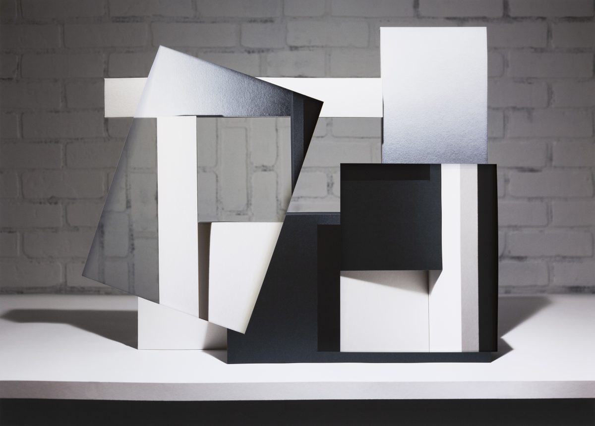 Abstract collage of white and black geometric forms layered and overlapped against a white brick wall backdrop. The composition features cut and folded paper planes creating sharp angles and shadows, with varied surface textures ranging from glossy to matte finishes, arranged in a three-dimensional sculptural arrangement on a white platform.