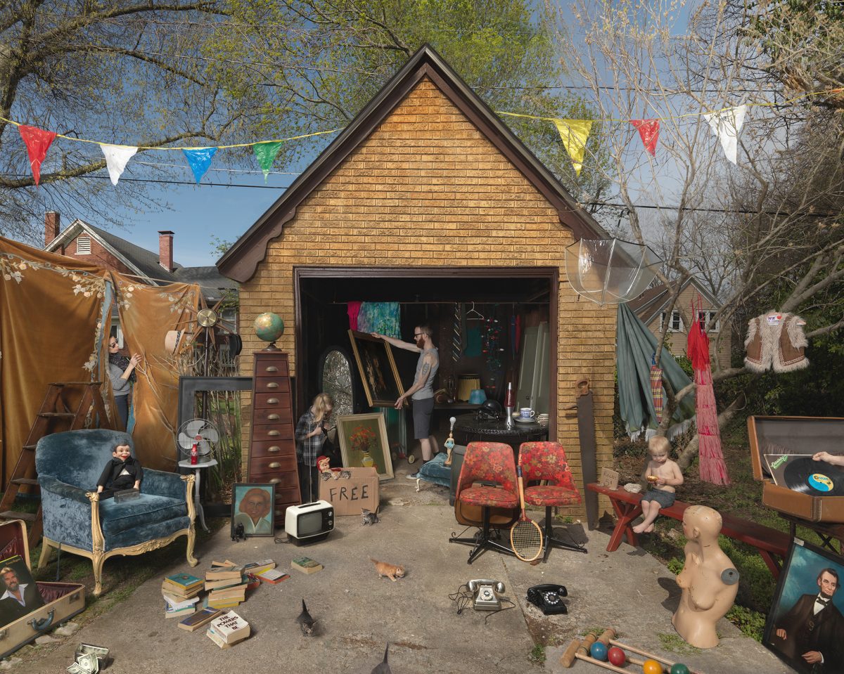 A suburban garage transformed into a bustling yard sale scene populated with mannequins and children among vintage furniture, artwork, textiles, and household goods. Colorful bunting flags string overhead while clothes hang from lines, creating a theatrical tableau of organized chaos and commerce.