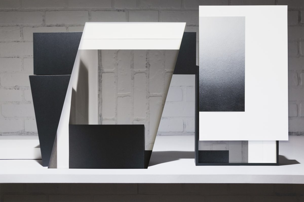 Abstract composition of collaged and layered white and black rectangular forms arranged on a white platform against a weathered brick wall, creating geometric planes that overlap and cast subtle shadows, with a minimalist aesthetic emphasizing contrasting values and spatial depth.