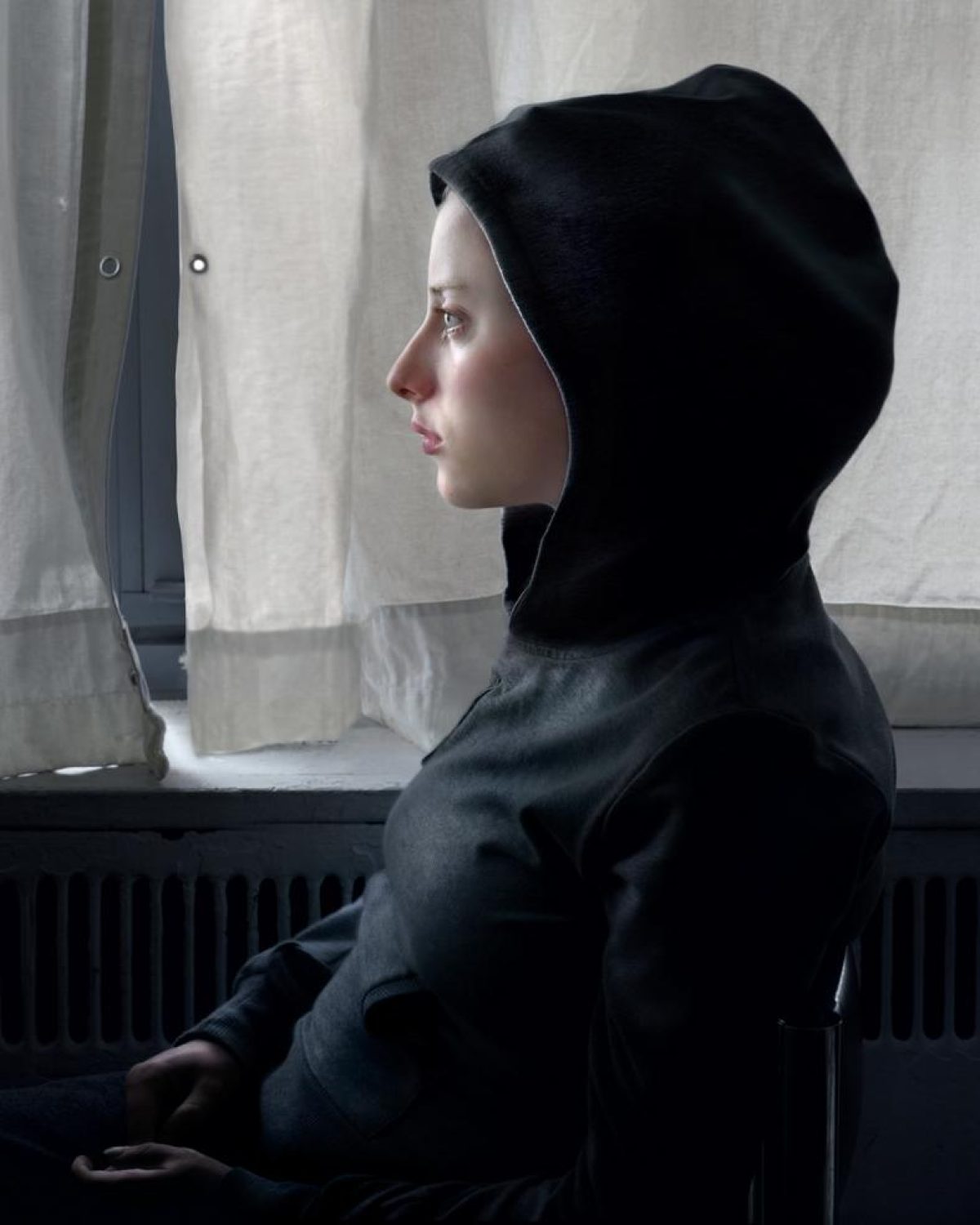 A figure in profile wearing a black hooded garment sits by a window with sheer white curtains. The minimalist composition features soft natural light against neutral tones and a dark radiator below the window sill.