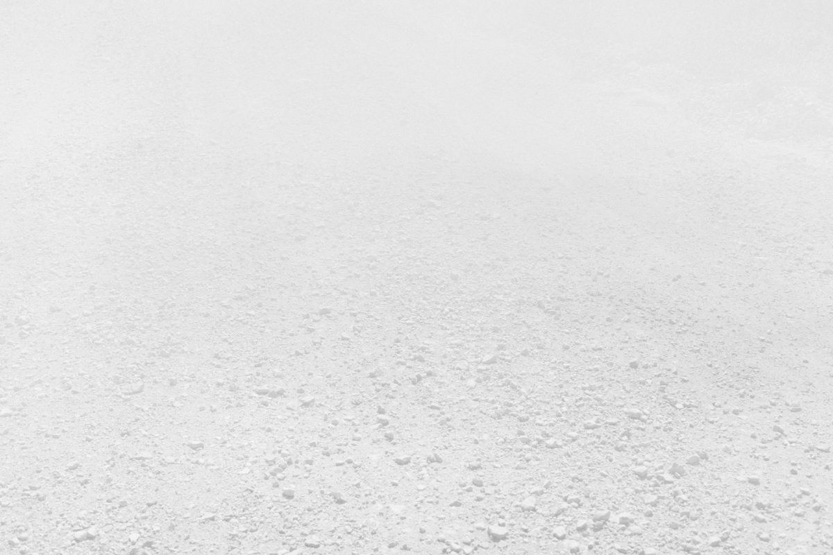 A vast, minimalist landscape rendered in pale gray and white tones, depicting an expansive flat terrain with subtle variations in texture and density across the surface. The composition recedes toward a hazy horizon, creating an immersive sense of emptiness and atmospheric perspective.