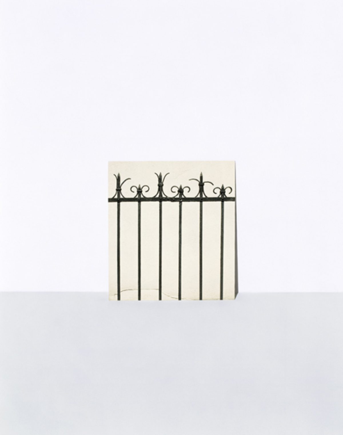 A minimalist composition featuring a decorative wrought iron fence with ornamental finials positioned against a pale cream-colored wall, casting sharp shadows on a light gray ground plane below.