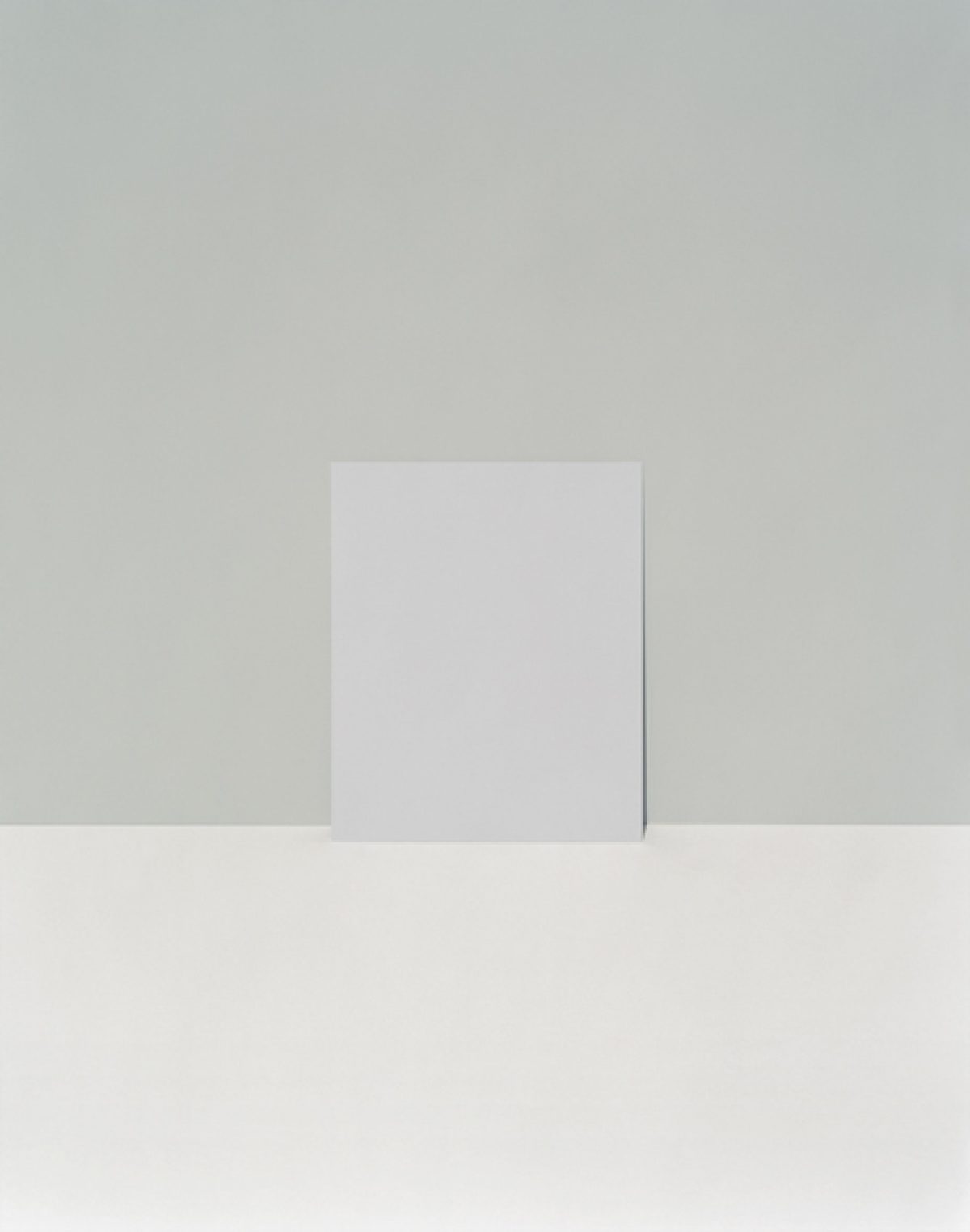 A minimalist composition featuring a white rectangular form standing vertically against a soft gray wall, casting a subtle shadow. The work employs a restrained palette of grays and whites with precise geometric forms, emphasizing negative space and the quiet interplay of light and shadow.