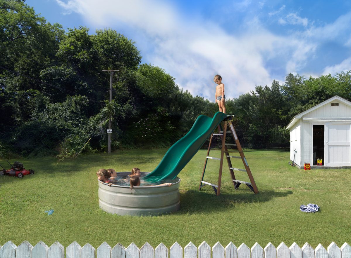Children play on a homemade water slide constructed from a green slide attached to a large metal stock tank in a residential backyard. One child stands atop the slide while others swim in the water below, surrounded by a manicured lawn, white picket fence, storage shed, and dense green trees under a bright blue sky.