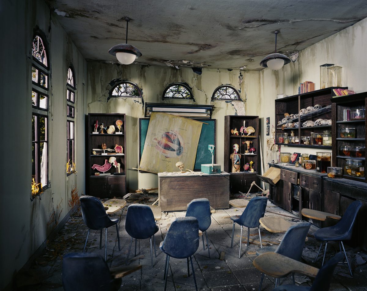 An abandoned classroom with decaying plaster walls and ceiling features modern blue chairs arranged around a wooden desk. Dark wood shelving units line the walls, displaying anatomical models, skulls, and educational materials beneath three arched windows. Anatomical diagrams and a skeleton figure are propped against the walls and desk, creating a haunting tableau of neglect and institutional decay.