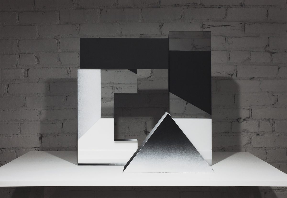 Abstract geometric composition featuring layered black and white rectangular forms arranged in an interlocking pattern against a weathered brick wall. A white triangular prism sits prominently in the foreground on a white platform, casting shadows that emphasize its three-dimensional form.