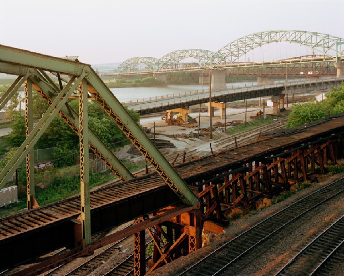 Industrial railroad bridge infrastructure in the foreground with rusted steel trusses and support beams, extending toward a modern arched bridge crossing a wide river in the distance. The composition layers deteriorating metalwork, active rail yards with equipment and structures, and waterway landscape under overcast sky, rendered in muted greens, browns, and grays.