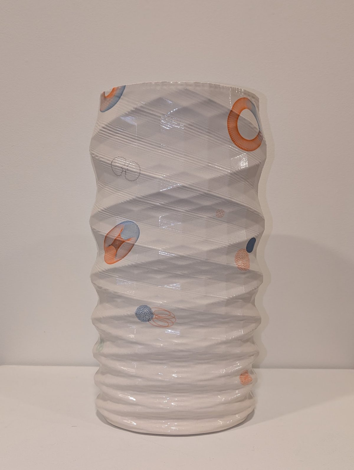A translucent cylindrical vessel with horizontal ridged texture, wrapped in diagonal bands of clear material. Small abstract geometric shapes in orange, blue, and coral tones are scattered across the surface, creating a sense of movement around the form.