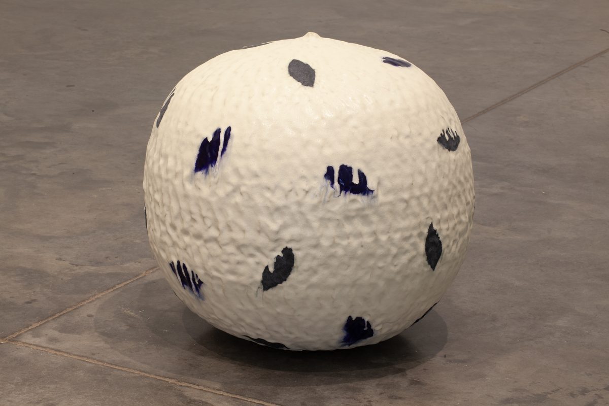 A large cream-colored spherical ceramic form sits on a concrete floor, its surface marked with gestural brushstrokes in deep blue and charcoal gray that resemble organic marks or leaf impressions distributed across the rounded form.