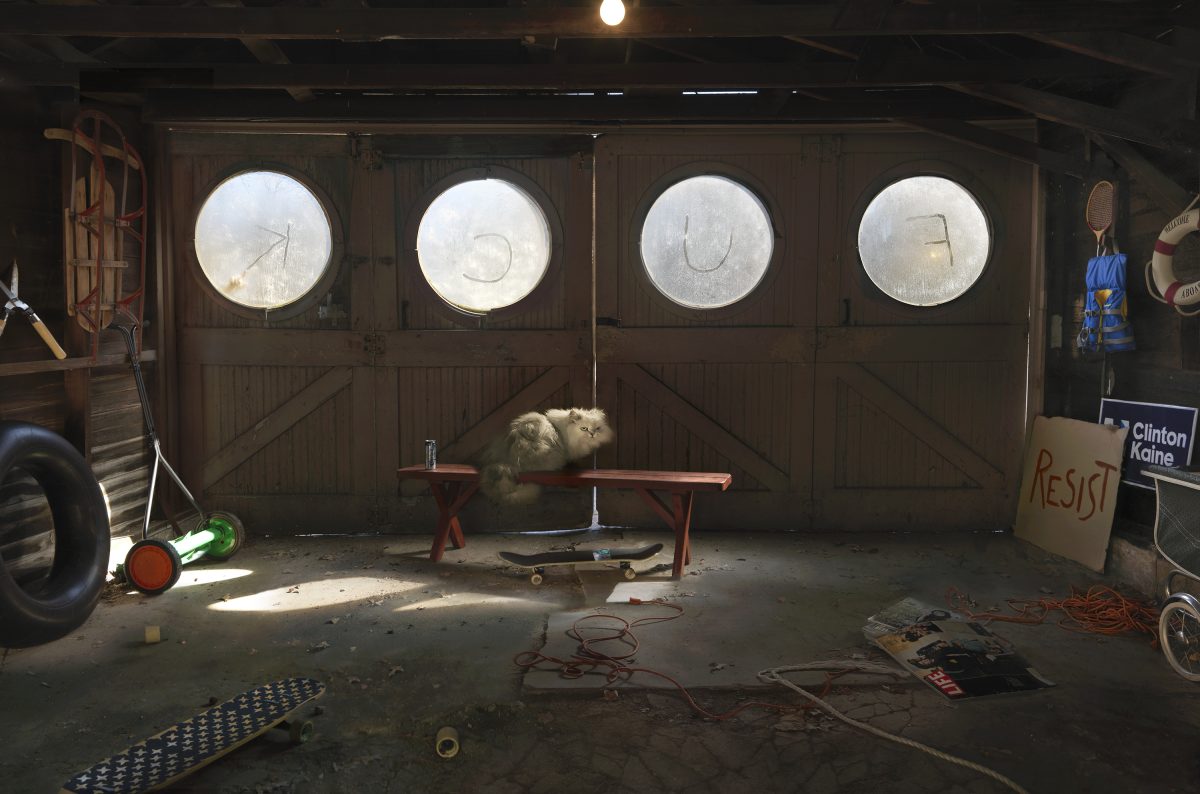 A cluttered garage interior featuring four large circular windows with frosted glass panes set into brown wooden doors. A fluffy white cat sits on a red bench beneath the windows. The concrete floor is scattered with miscellaneous objects including bicycle parts, coiled red cable, a skateboard, sports equipment, and political signage. Sunlight streams through the windows, creating bright geometric patterns across the space.