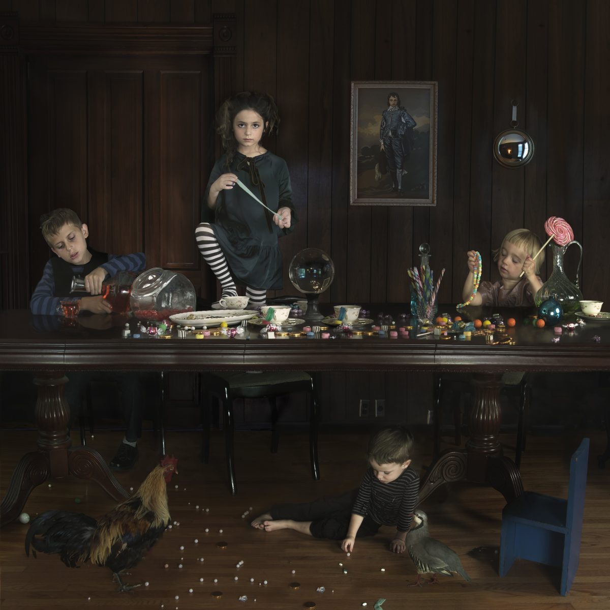 Children of varying ages are arranged at a long dark table laden with candies and toys, with two younger children and a rooster on the floor below scattering small round objects. The scene is set in a wood-paneled interior with framed portraits on the walls, rendered in rich, dark tones with carefully controlled lighting that emphasizes the children's faces and the colorful items scattered across the surfaces.
