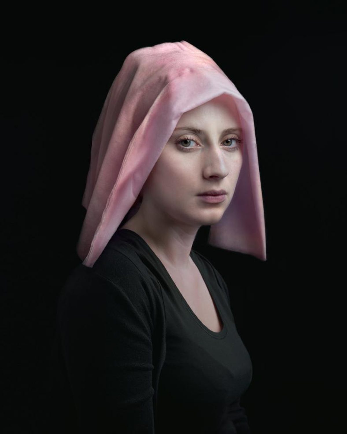 Portrait of a young woman in profile against a black background, wearing a black long-sleeved top and a draped pink fabric covering her head. Her pale complexion and direct gaze create a contemplative mood, with soft lighting emphasizing the delicate folds of the translucent cloth.