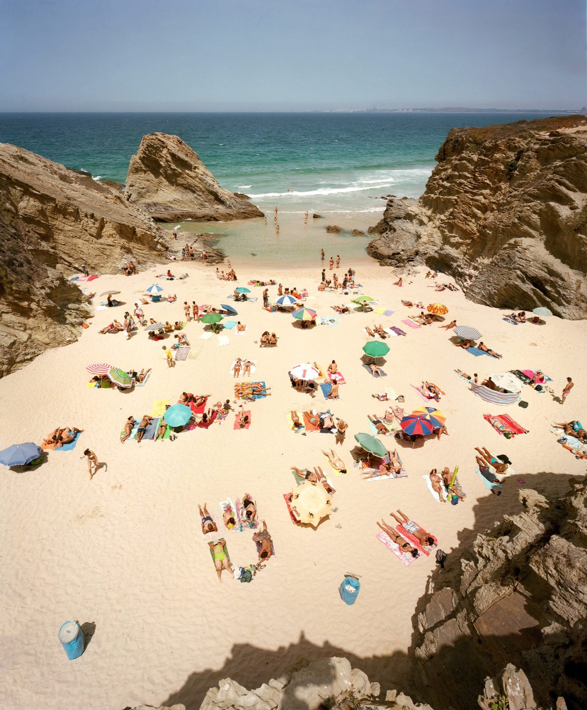 Aerial view of a crowded beach cove framed by dramatic rocky outcrops, with dozens of beachgoers scattered across pale sand beneath colorful umbrellas and towels, turquoise water visible beyond the shore.