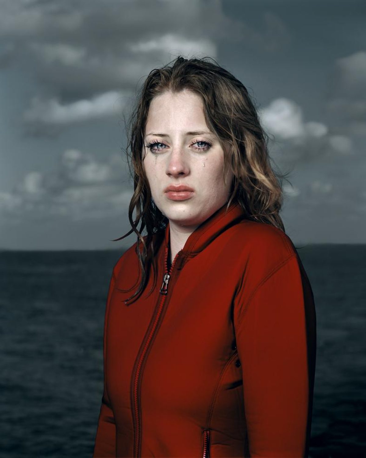 Portrait of a woman with dark, wet-looking hair wearing a bright red zip-up jacket, photographed against a stormy seascape with overcast sky and turbulent water.