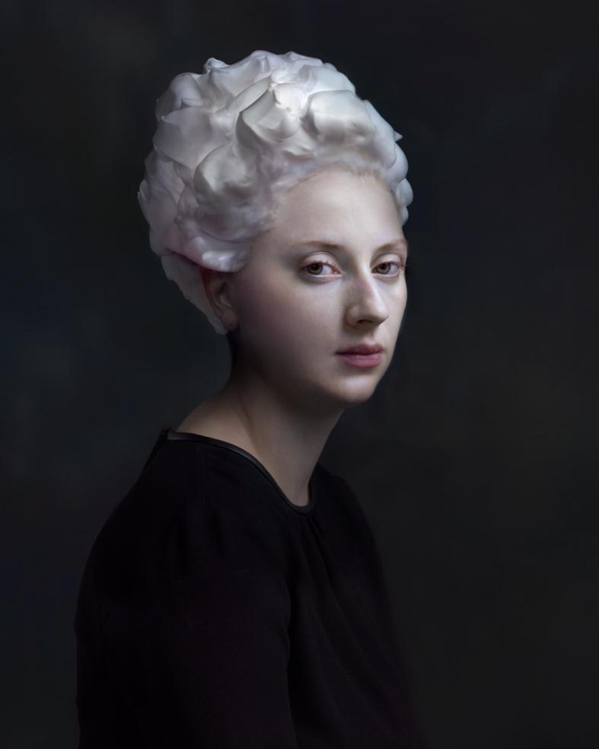 Portrait of a person with pale skin and white sculptural hair styled in voluminous waves, wearing a black garment against a dark background. The lighting is soft and dramatic, emphasizing the figure's serene expression and the luminous quality of the hair.