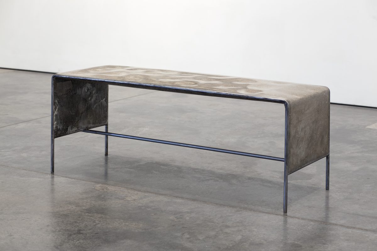 Industrial bench with a concrete and ceramic top featuring geometric surface patterns, supported by dark metal legs and frame with blue-tinted steel accents. The weathered materials and minimalist construction create a stark, contemporary sculptural form displayed on polished concrete flooring.