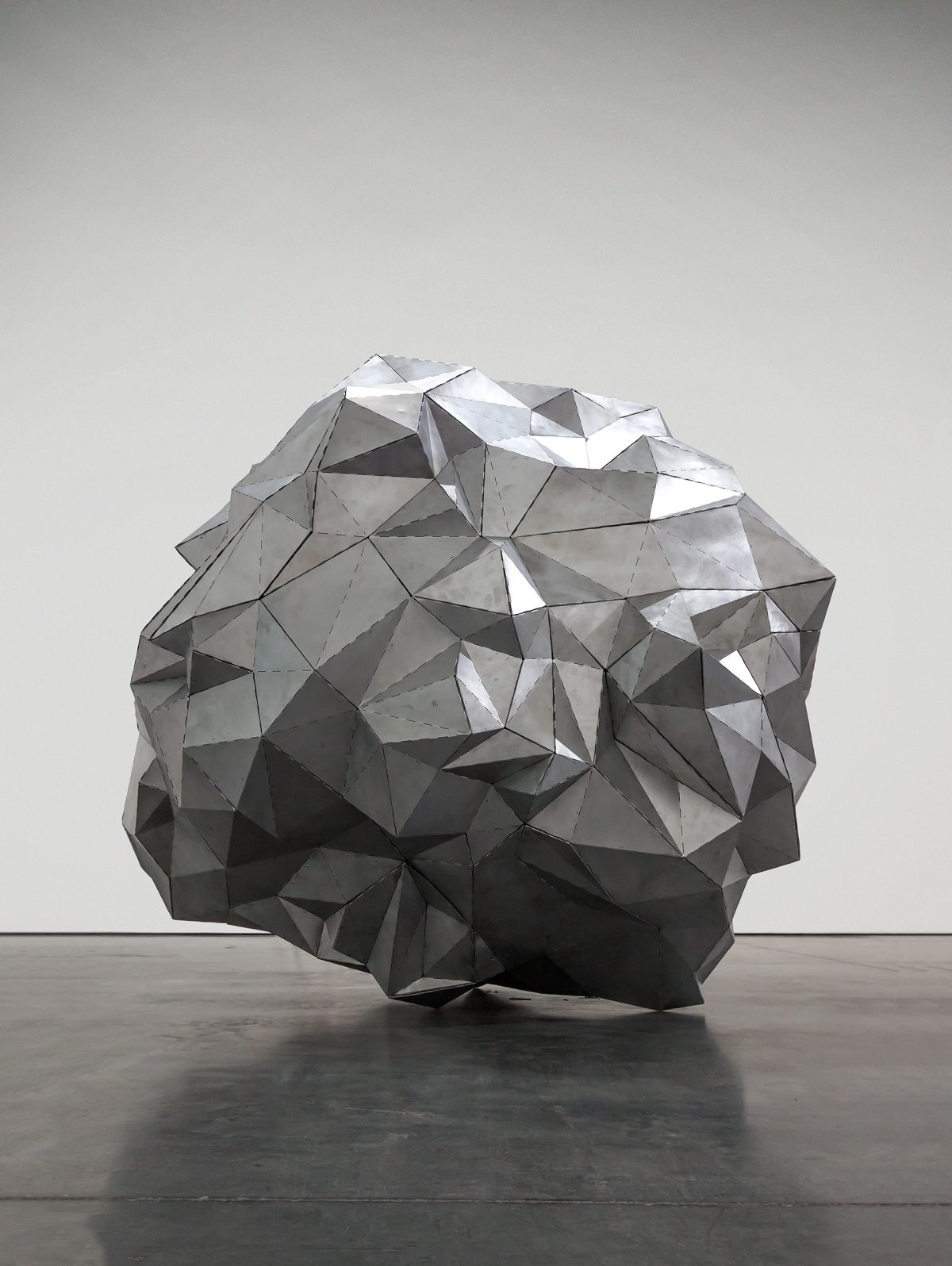 A large spherical sculpture composed of folded aluminum planes creates a faceted, geometric surface. The form is rendered in shades of gray and white, with dramatic shadows cast by the angular facets, set against a neutral gallery wall and polished concrete floor.