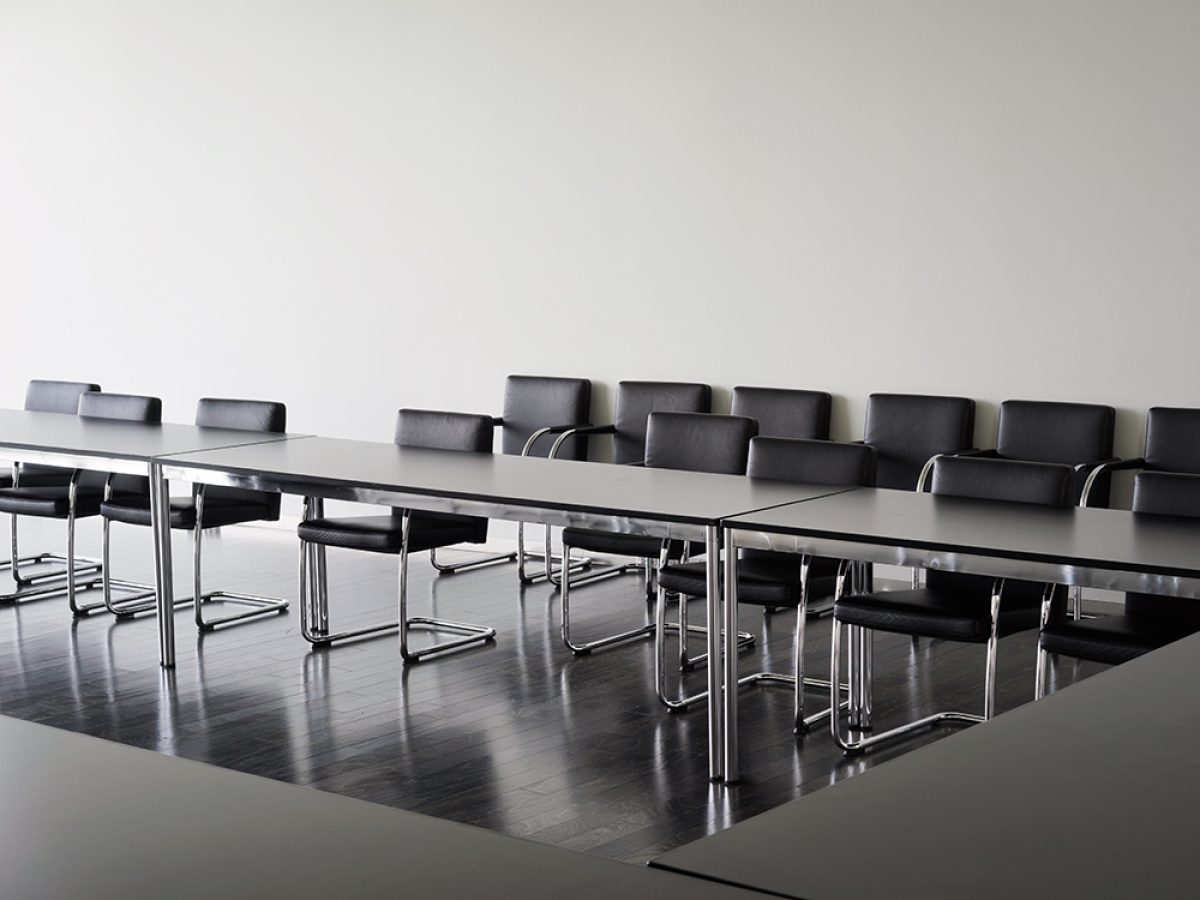 A minimalist interior photograph depicting a sleek conference room with a long, reflective dark table surrounded by black leather chairs with chrome frames. The polished dark flooring mirrors the table's surface, while white walls and subtle overhead lighting create a stark, contemporary aesthetic emphasizing geometric forms and material contrasts.