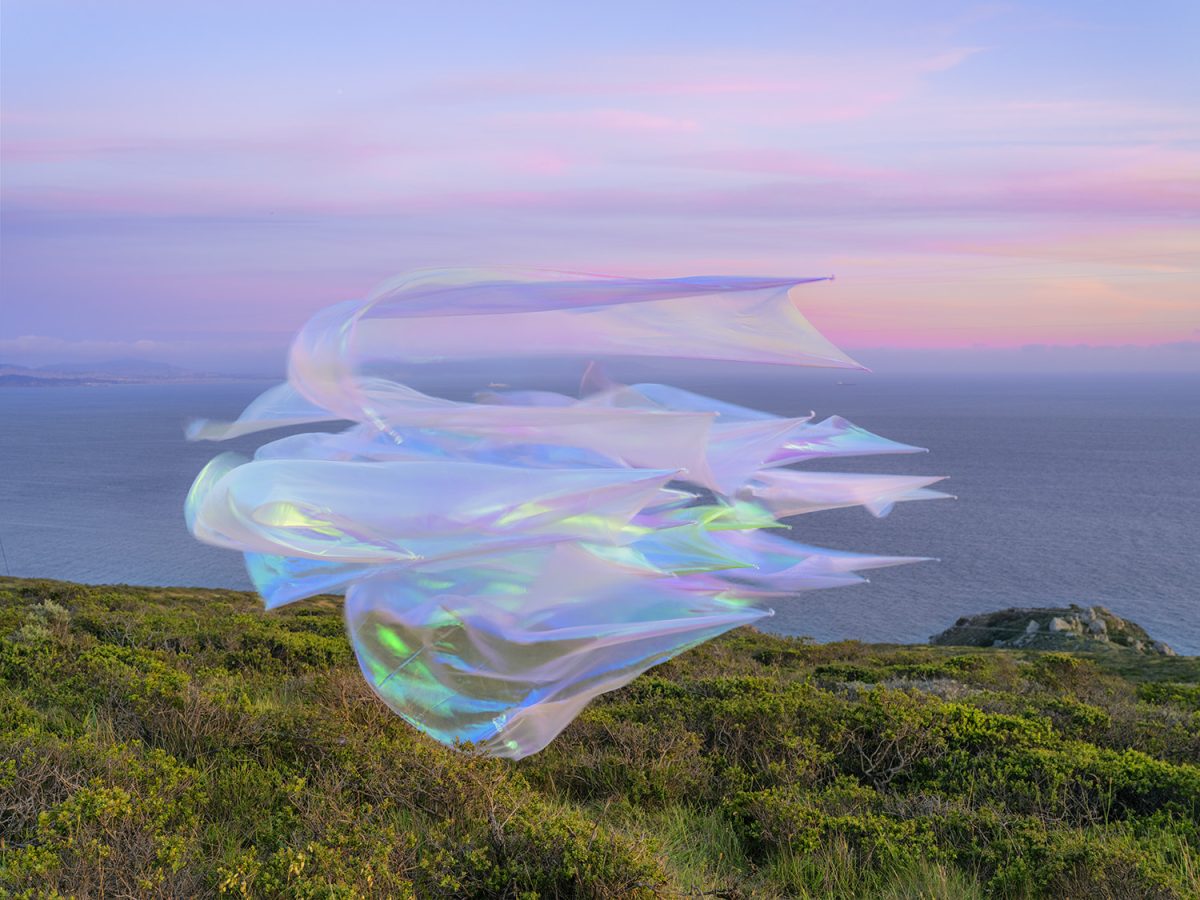 Iridescent, translucent fabric forms hover above a coastal hillside overlooking the ocean at dusk, their flowing surfaces catching pink, blue, and green light against the soft twilight sky.