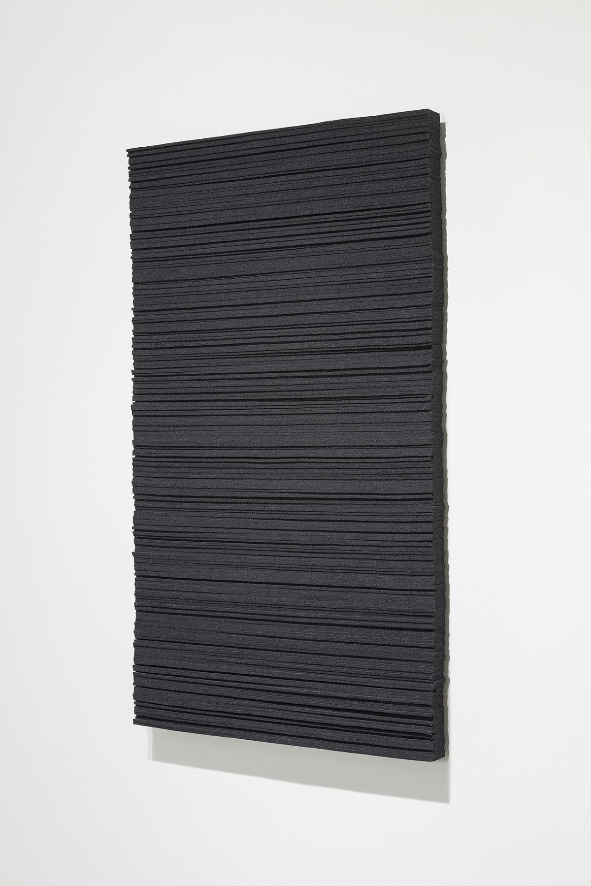 A rectangular panel of dark charcoal wool felt with horizontal linear ridges across its surface, mounted at a slight angle against a light gray background with a pale base visible beneath.