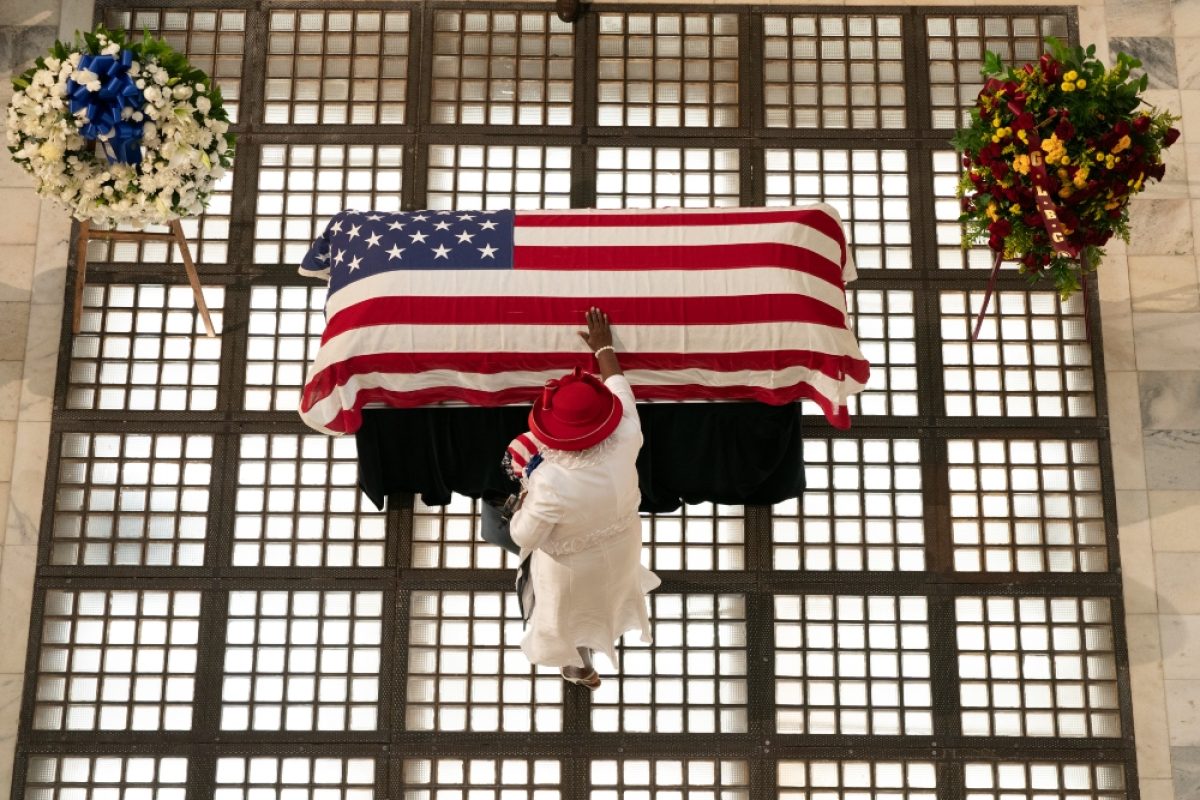 Overhead view of a person in white clothing and a red hat standing before a casket draped with the American flag, flanked by two floral wreaths on either side.