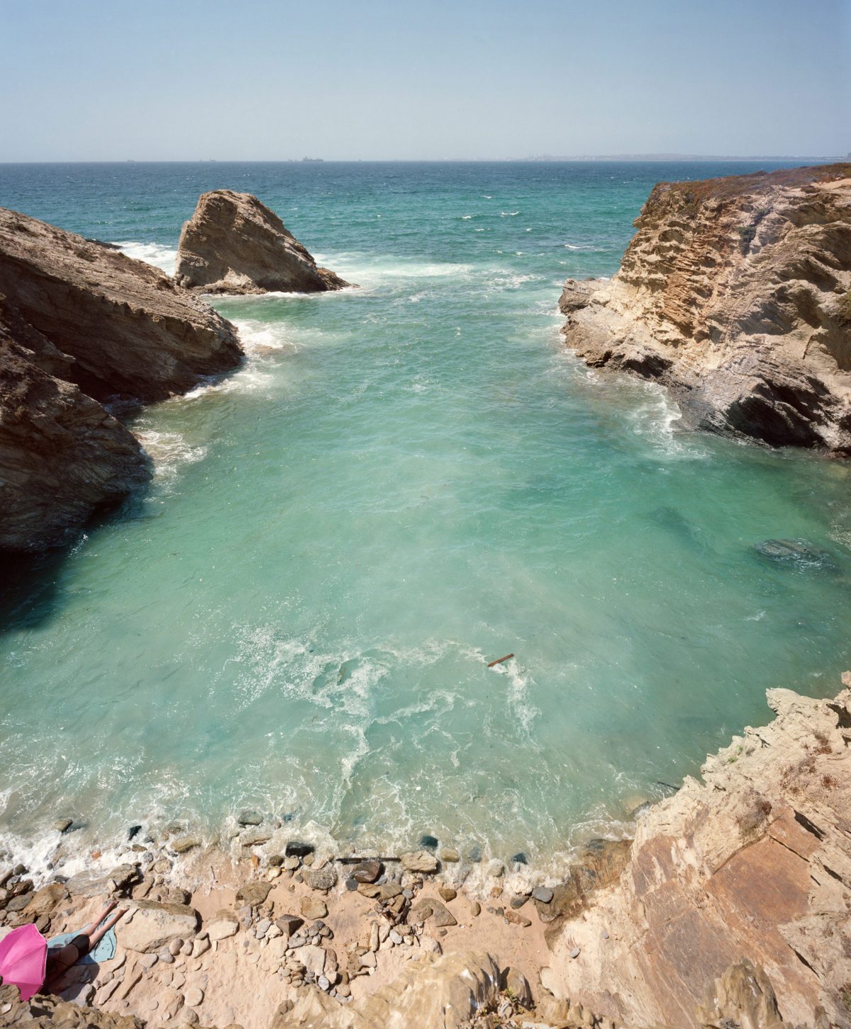 Coastal landscape featuring turquoise waters framed by rocky limestone cliffs, with a solitary rock formation rising from the sea and a small sandy beach in the foreground where a person in pink clothing sits.