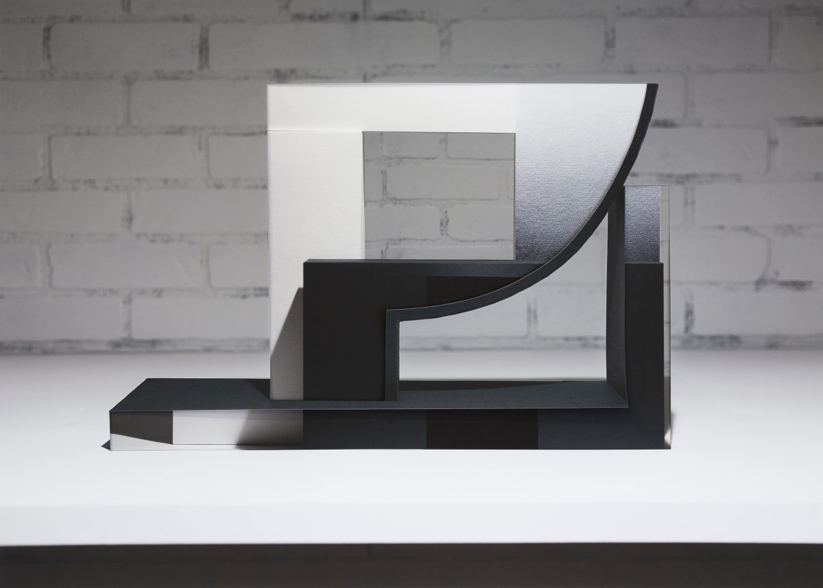 Abstract black sculptural form composed of geometric planes and curved elements, displayed against a white gallery wall with visible brick texture. The composition features layered, translucent paper elements in white and gray tones creating depth and dimensionality through collage and decollage techniques.
